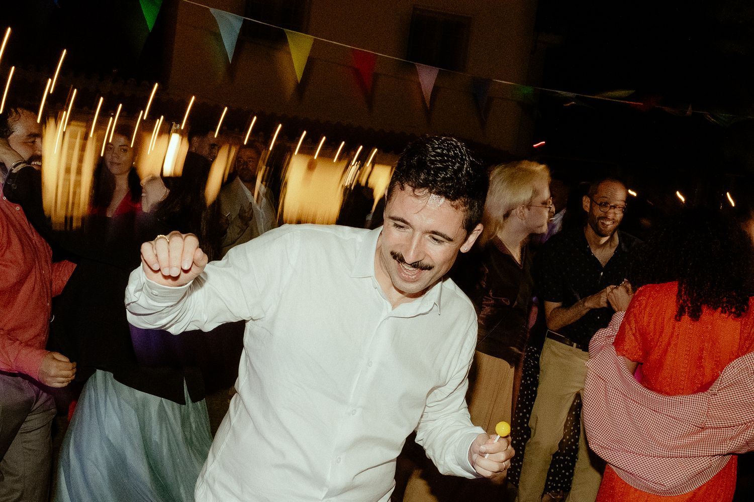 Man with mustache dancing energetically at an outdoor night party with colorful bunting flags in background.
