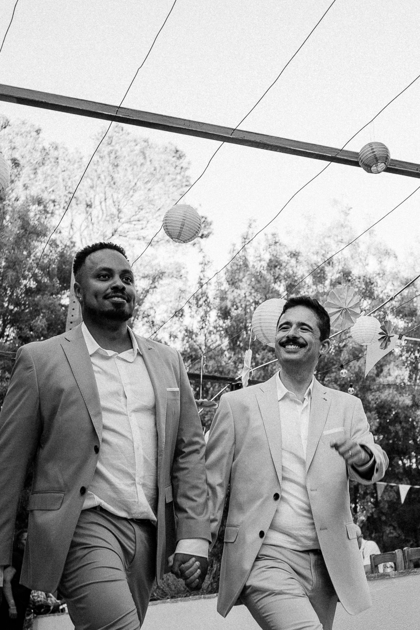 Two grooms in matching suits hold hands and smile while walking under paper lantern decorations at their outdoor wedding.