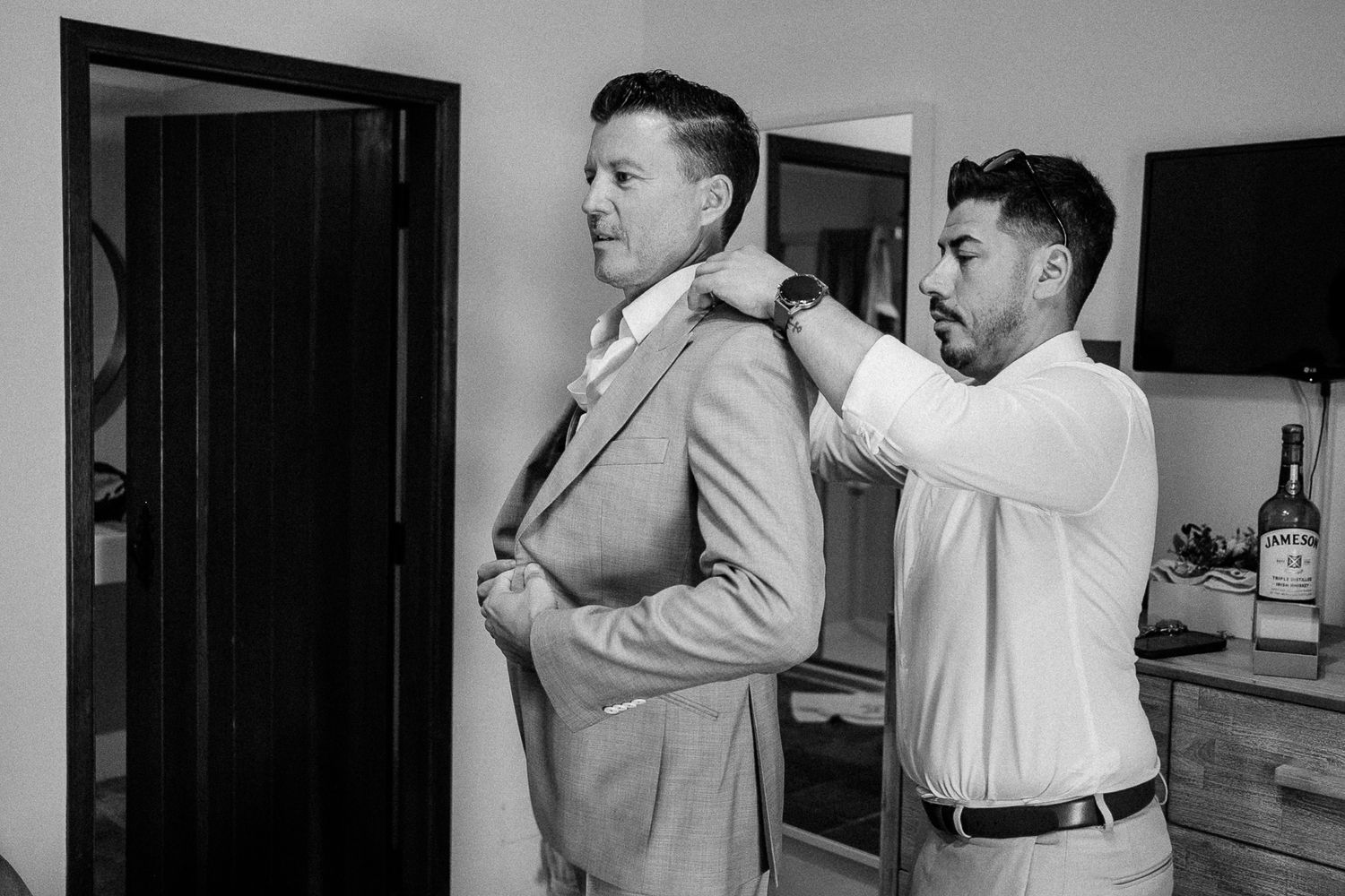 Black and white photo of a man helping a groom adjust his suit jacket before a wedding ceremony.