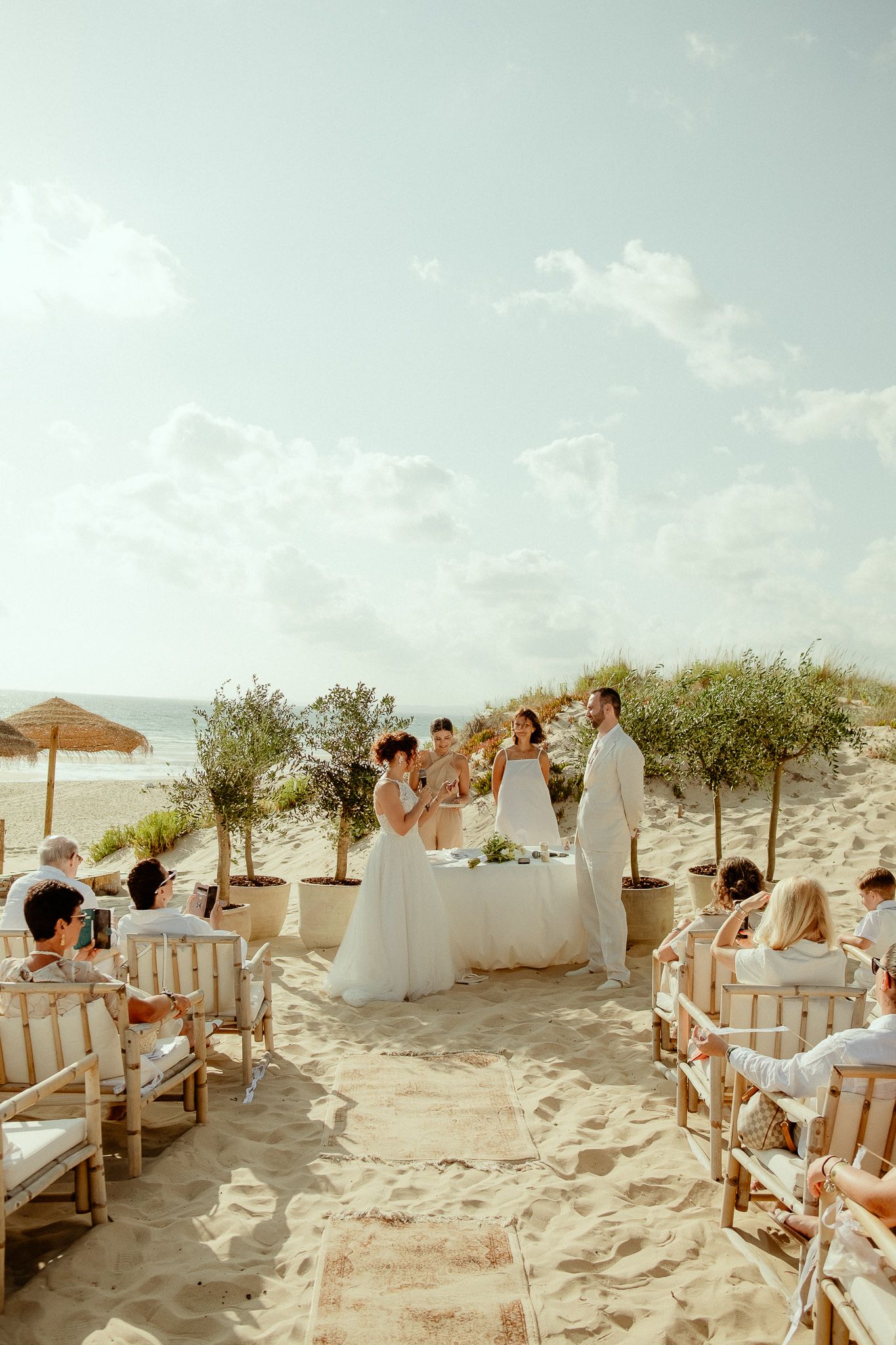 Romantic beach wedding ceremony with bride and groom at altar, guests seated on wooden chairs in sandy coastal setting.