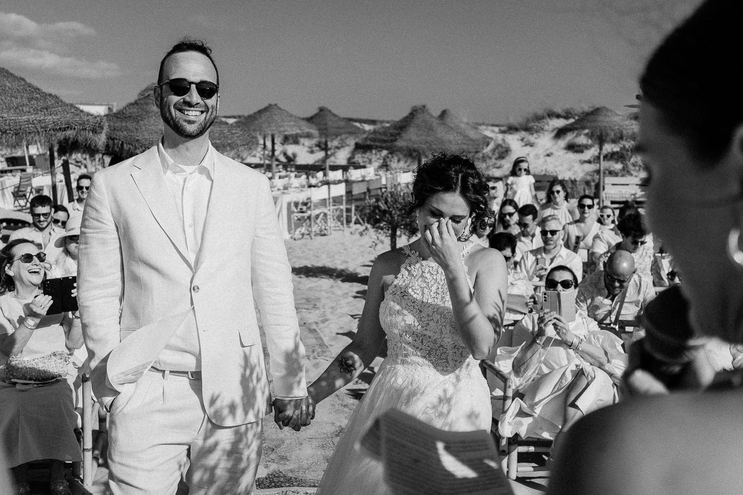 Bride and groom hold hands smiling at outdoor beach wedding ceremony with guests in background.