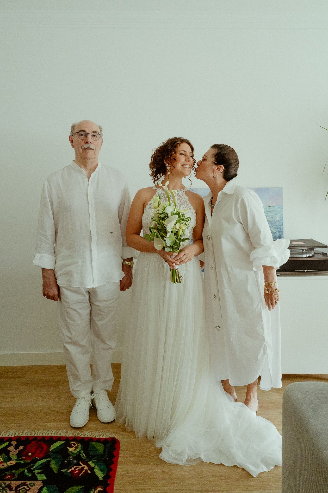 Bride in white tulle gown holds green bouquet while parents kiss her cheek indoors before wedding ceremony.