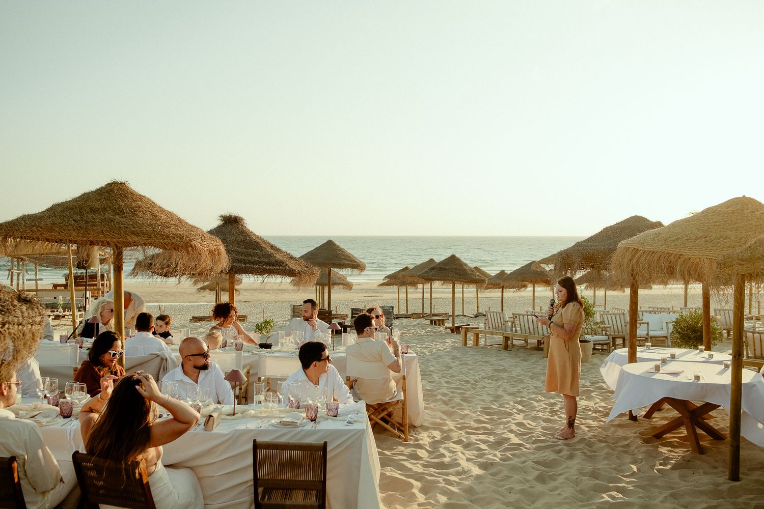 Elegant beachfront dining with white tablecloths, thatched umbrellas, and guests enjoying a meal on sandy shores at sunset.