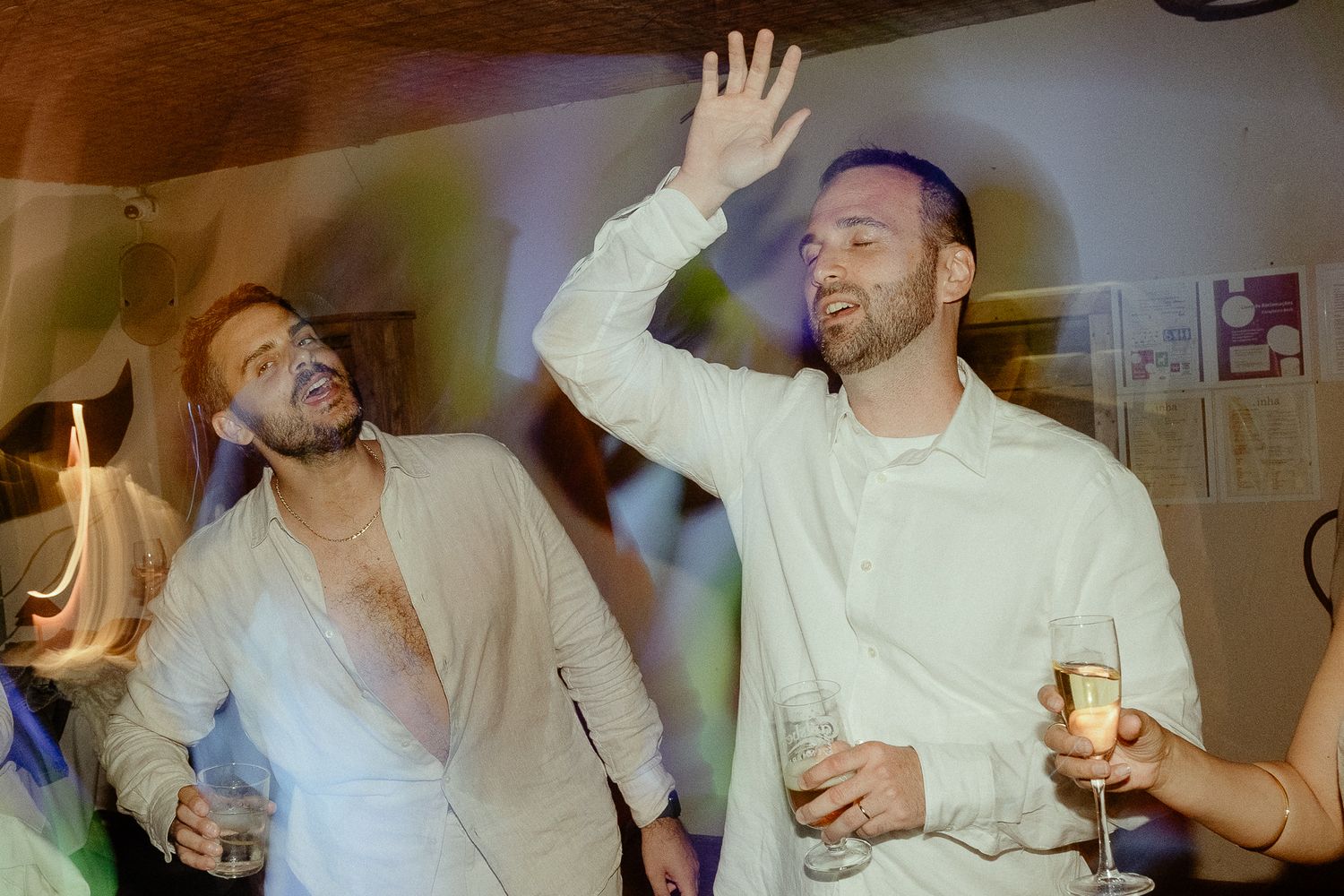 Two men in white shirts celebrate at a party, one raising his hand while holding a champagne glass.
