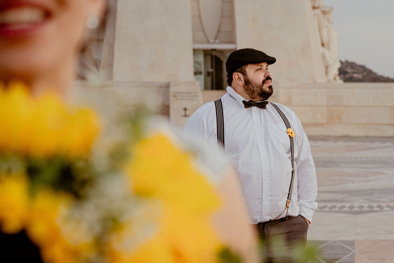 People in formal attire gathering outdoors at a stone building with yellow flowers in foreground.