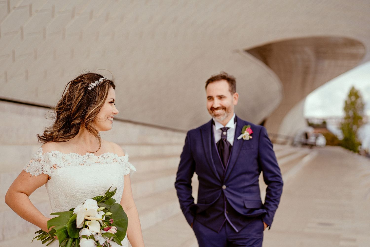 Wedding couple stands under modern architectural arch with bride holding white bouquet.