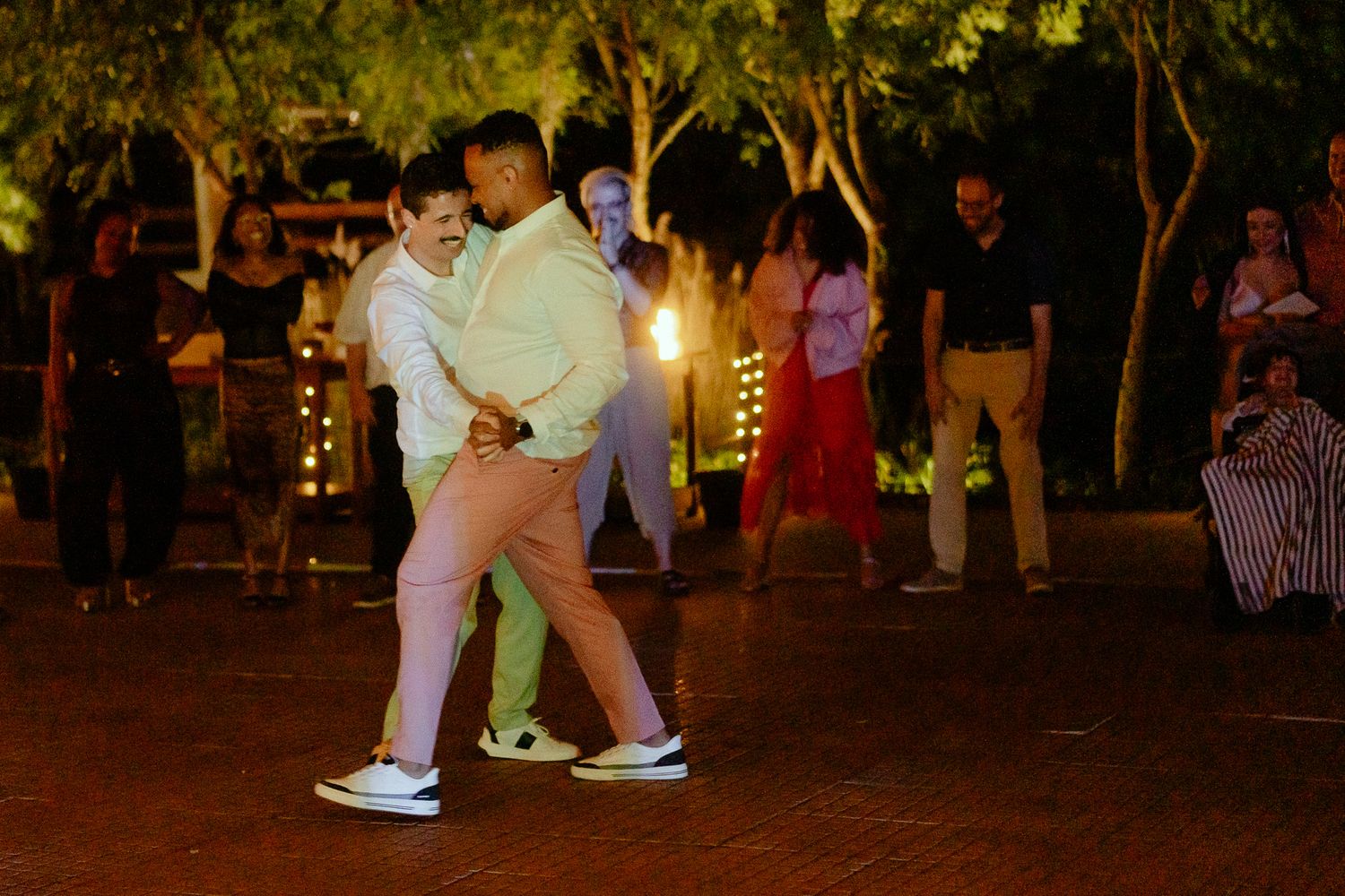 Two men dancing together outdoors at a nighttime event on a brick patio with trees and guests in background.