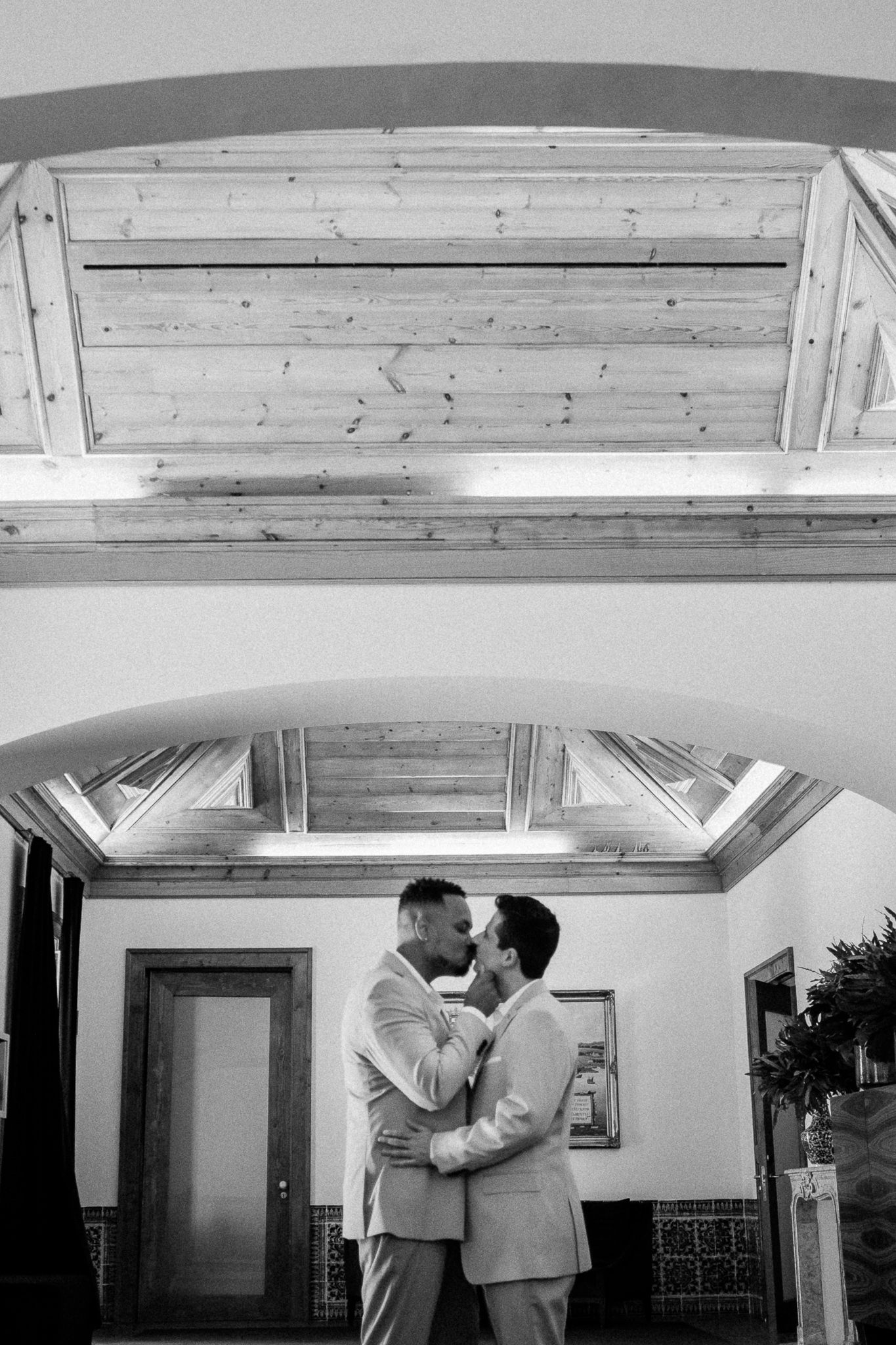 Two grooms share a romantic embrace in black and white wedding photo beneath elegant wooden ceiling.