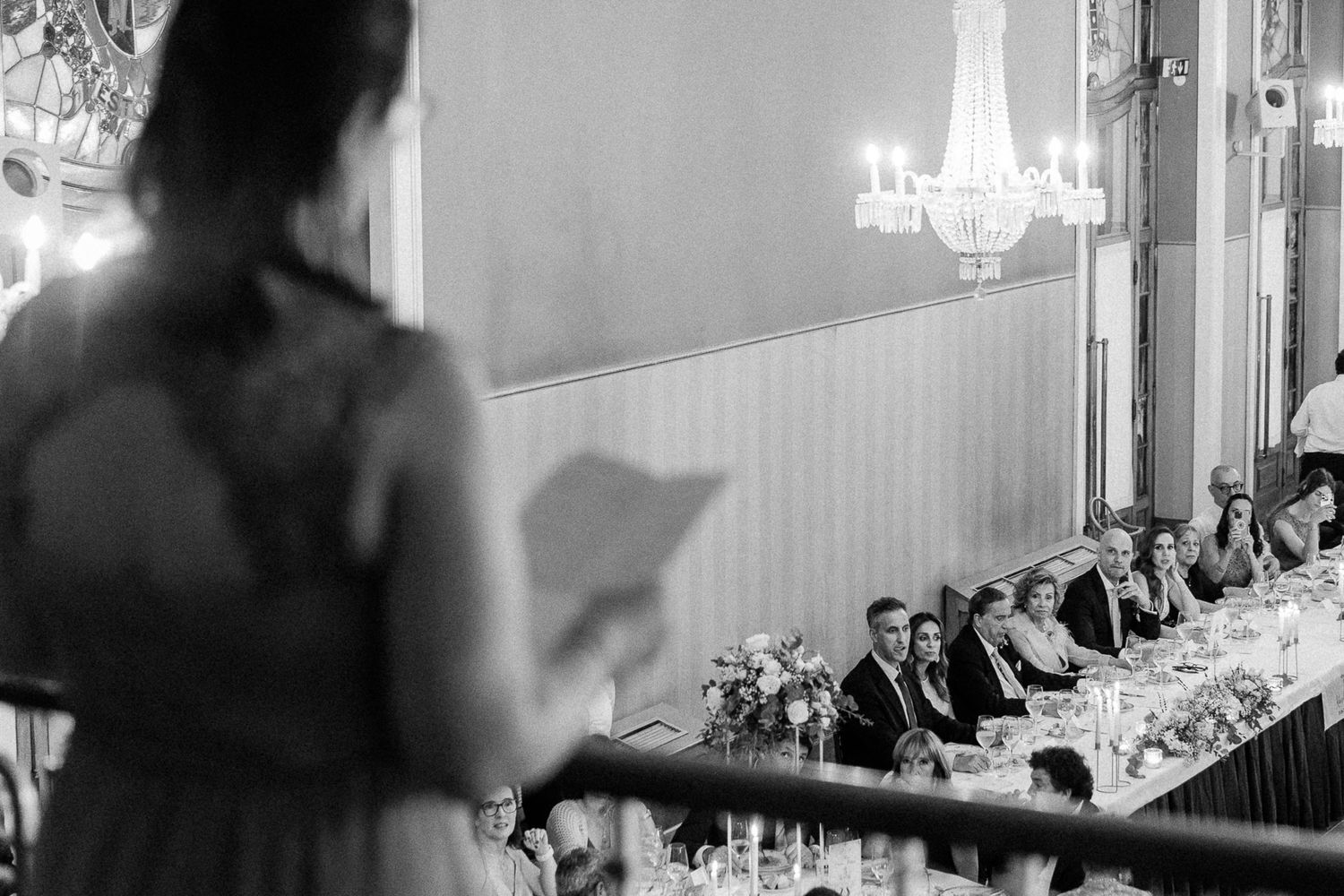 Black and white wedding reception photo of a woman giving a speech at an elegant venue with chandeliers.