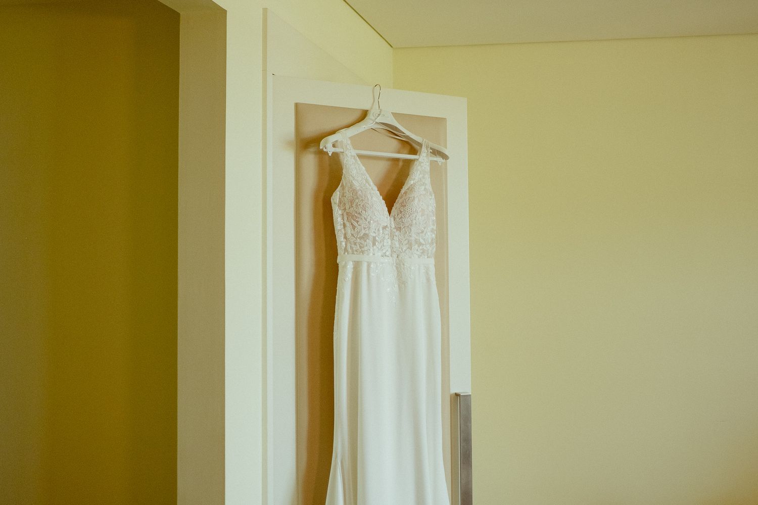 Elegant white wedding dress with lace bodice and V-neckline hanging on a door hanger in a softly lit room.