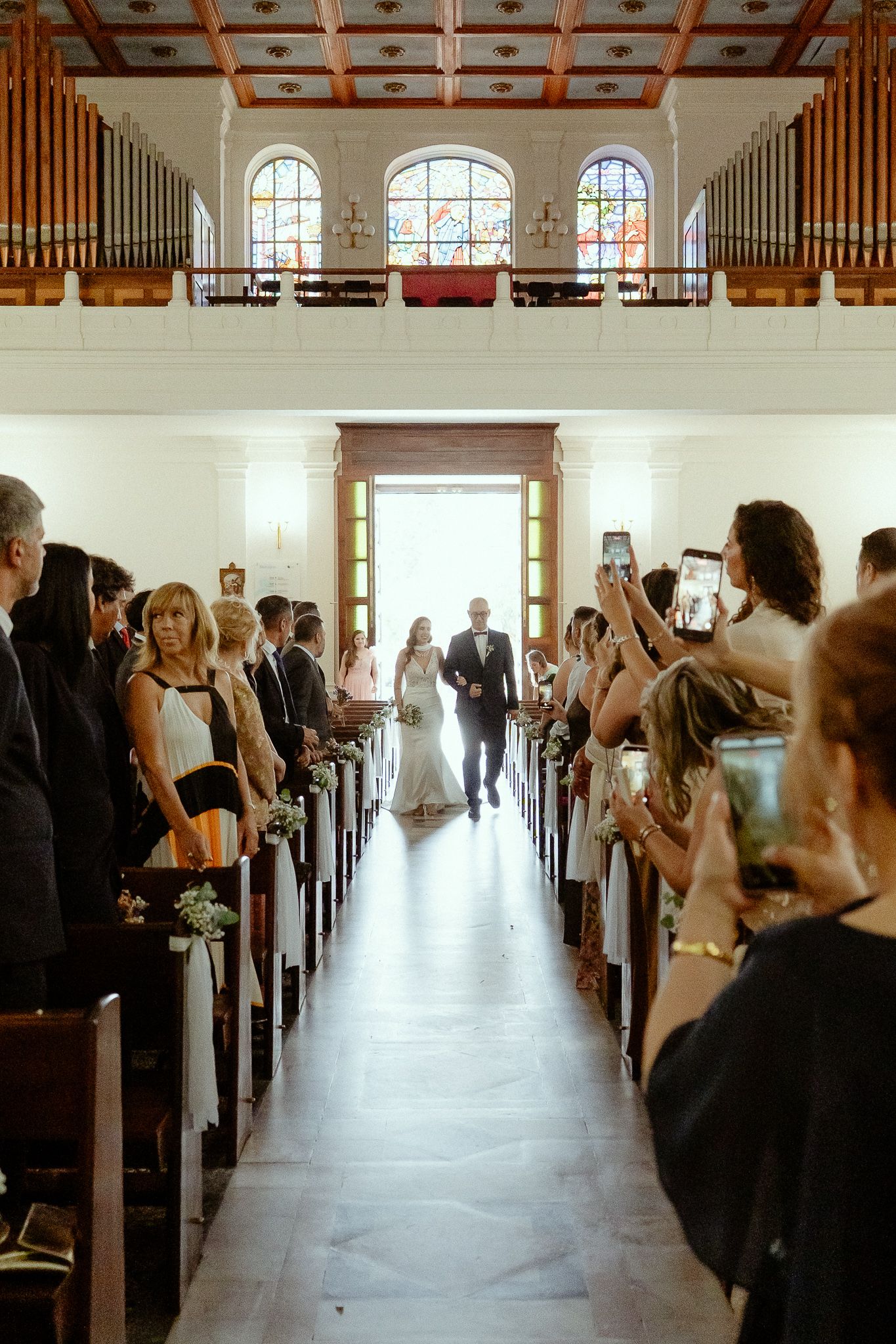 Bride walking down the aisle with her father in a sunlit church, guests capturing the moment on phones.