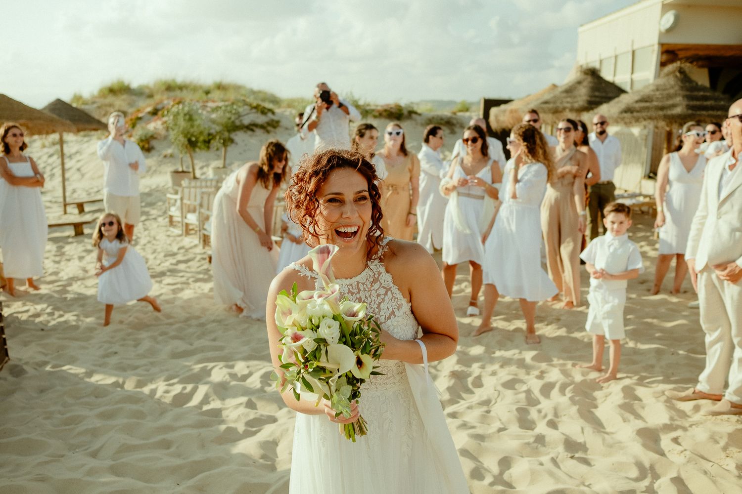 Bride holding bouquet smiles on sandy beach with wedding guests in white attire gathered behind her.