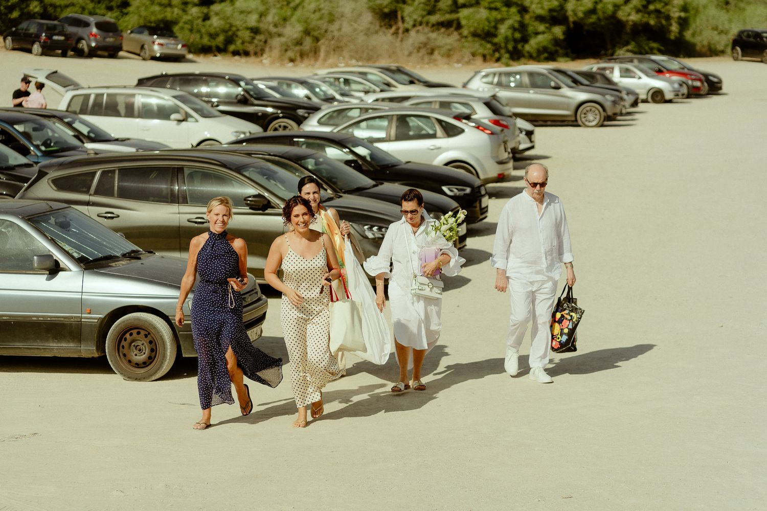 Group of friends walking through a sunny parking lot, dressed in summer attire, carrying bags and flowers.