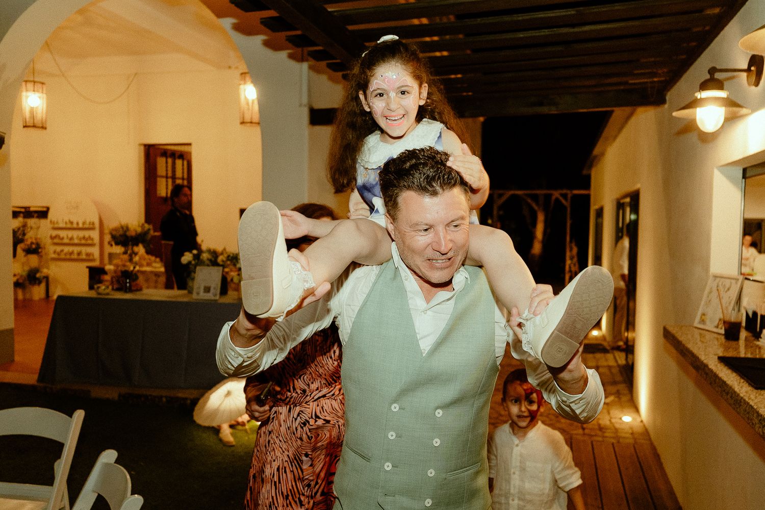 Man in mint vest carries smiling girl on shoulders at elegant indoor wedding reception celebration.