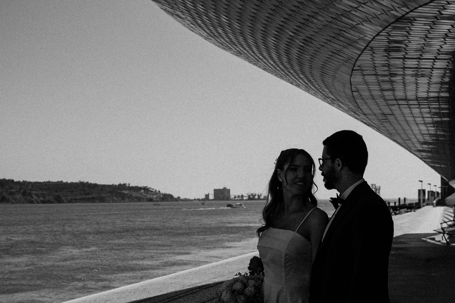 Black and white photo of a bride and groom sharing a romantic moment by a waterfront under a modern architectural canopy.