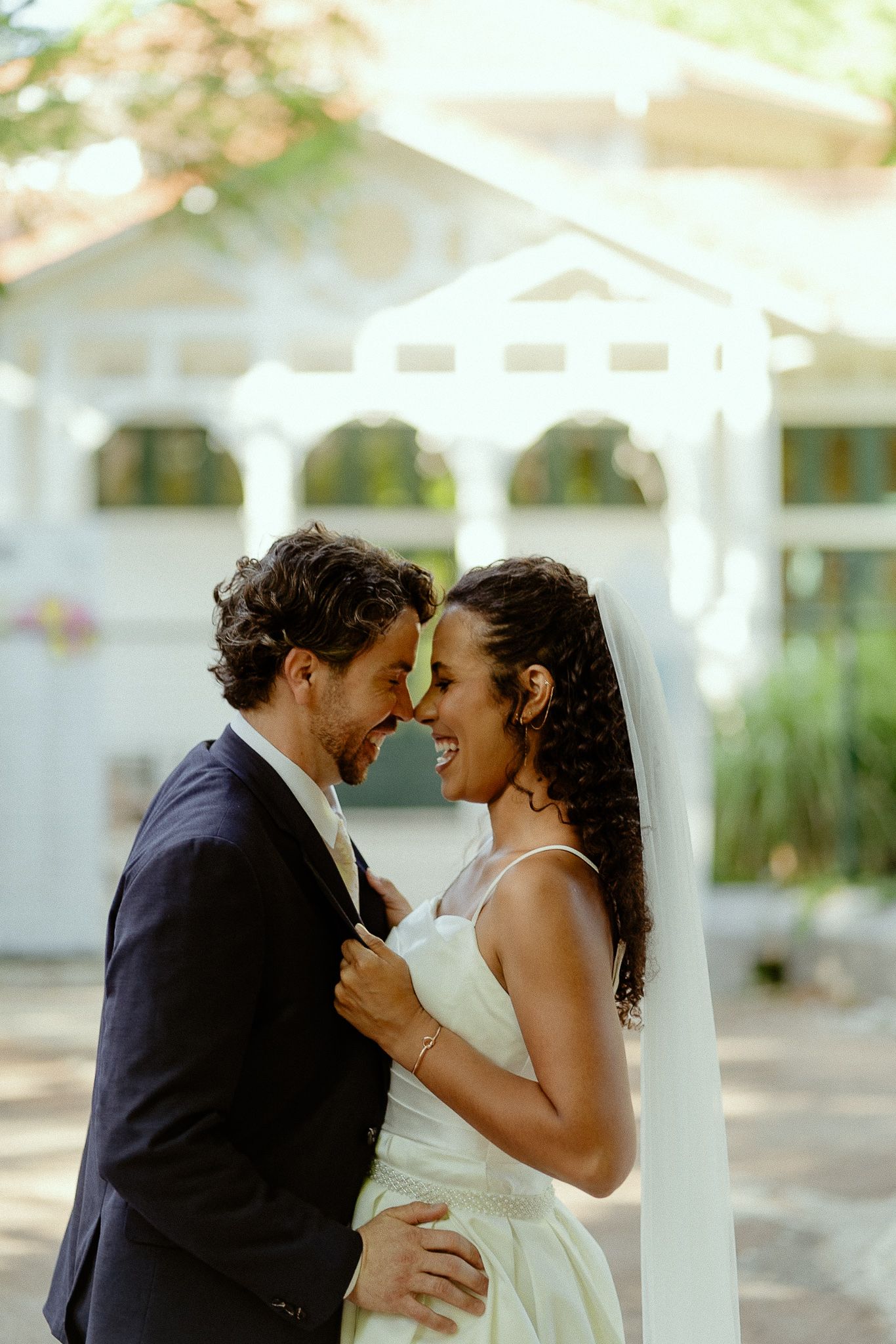A joyful interracial couple shares a romantic moment in wedding attire outdoors near a white gazebo.