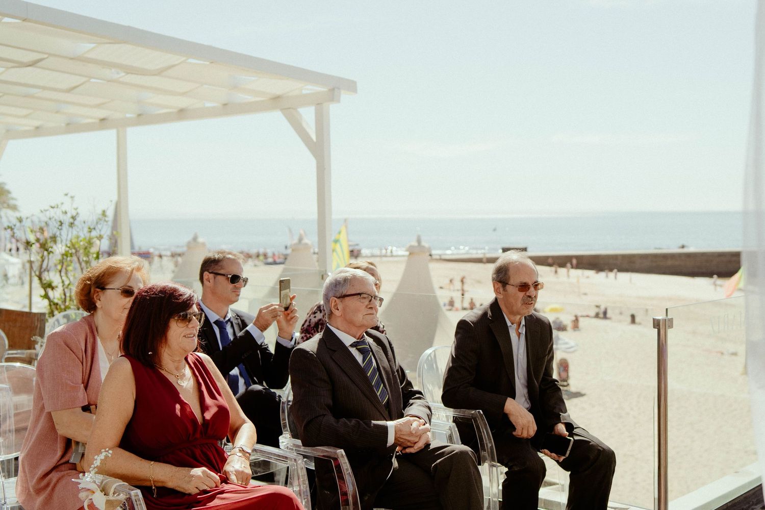 A group of people sitting on a bench at a beach overlook with a telescope viewer on a sunny day.