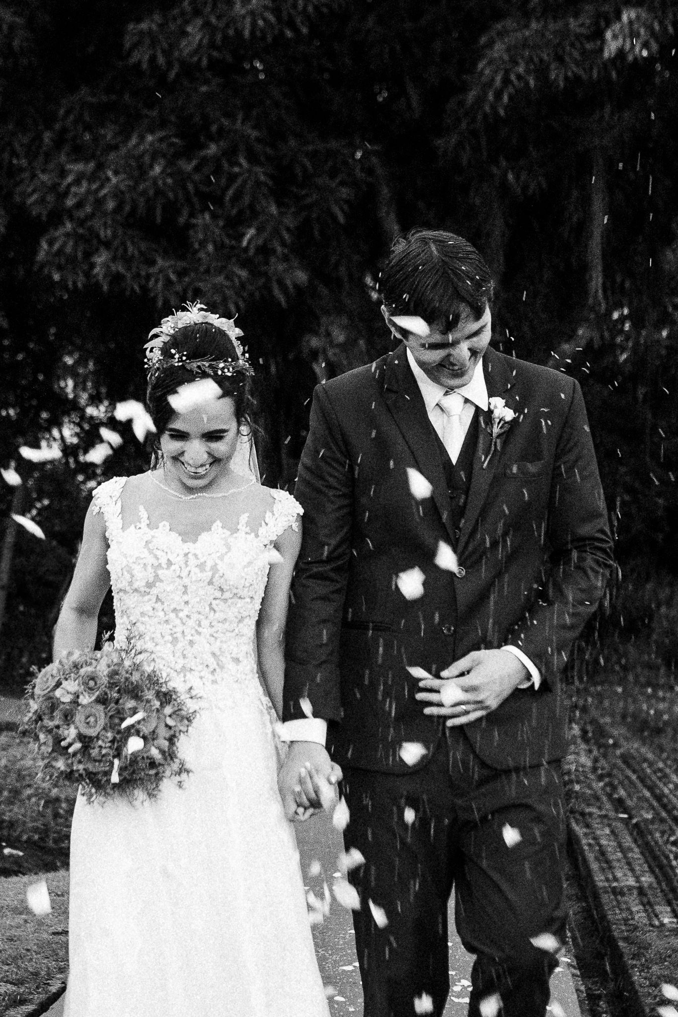 A happy couple walks together in formal attire while white flower petals fall around them during an outdoor celebration.