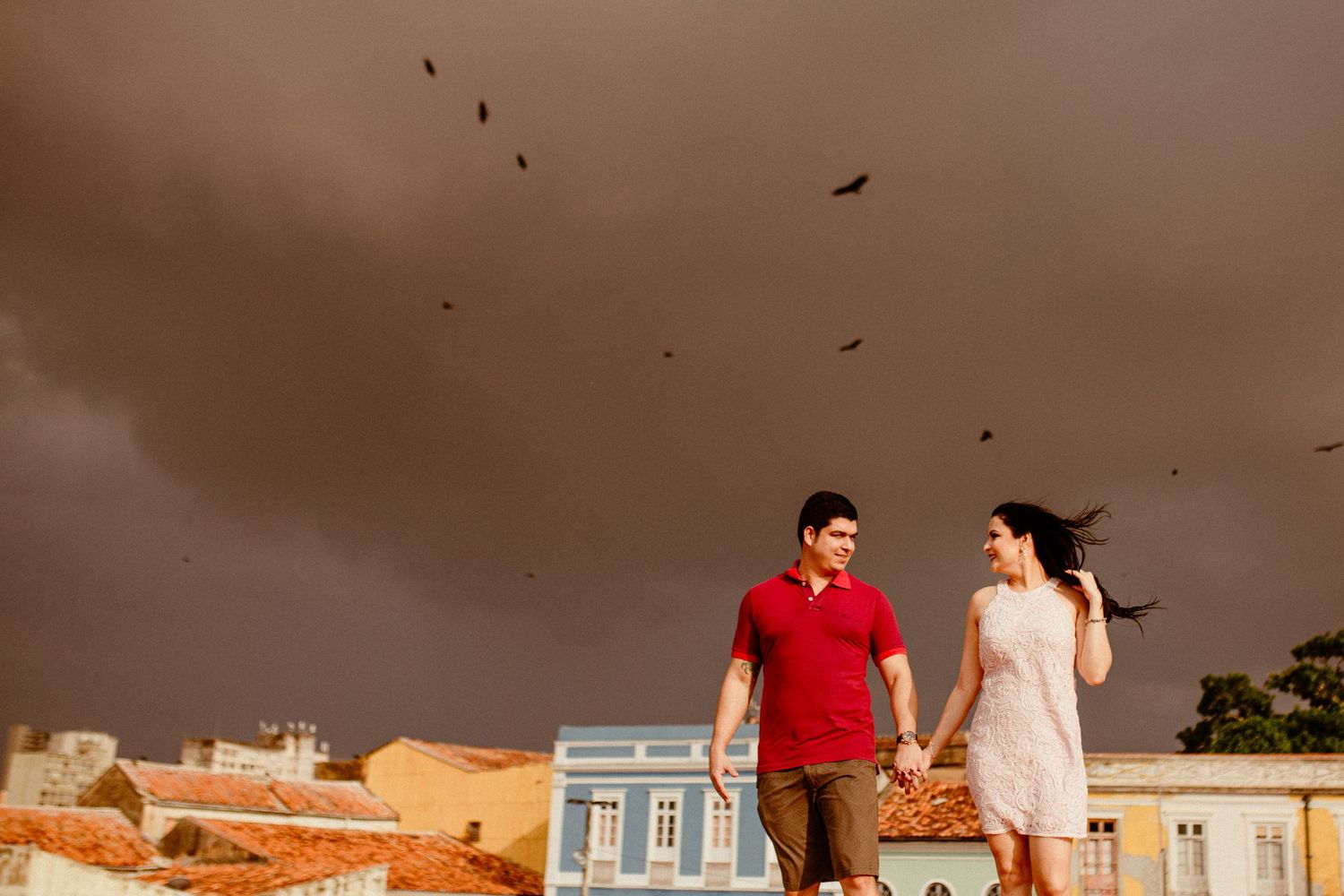 A couple stands on rooftops at sunset with birds flying overhead and colorful buildings in the background.