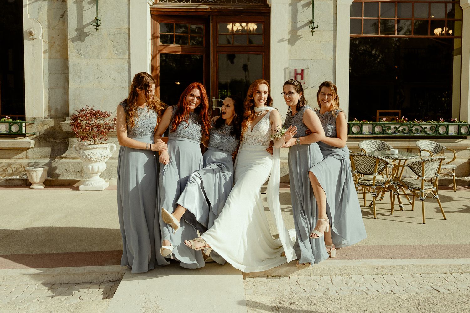 Bride in white gown poses with four bridesmaids in dusty blue dresses outside elegant venue.