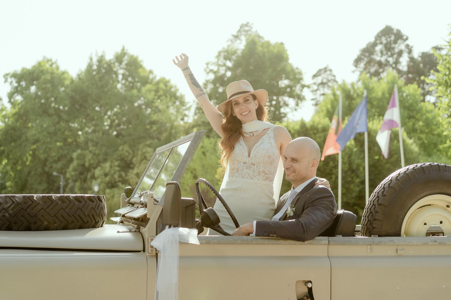 Bride and groom riding in an open-top vintage jeep outdoors, woman waving joyfully on their wedding day.