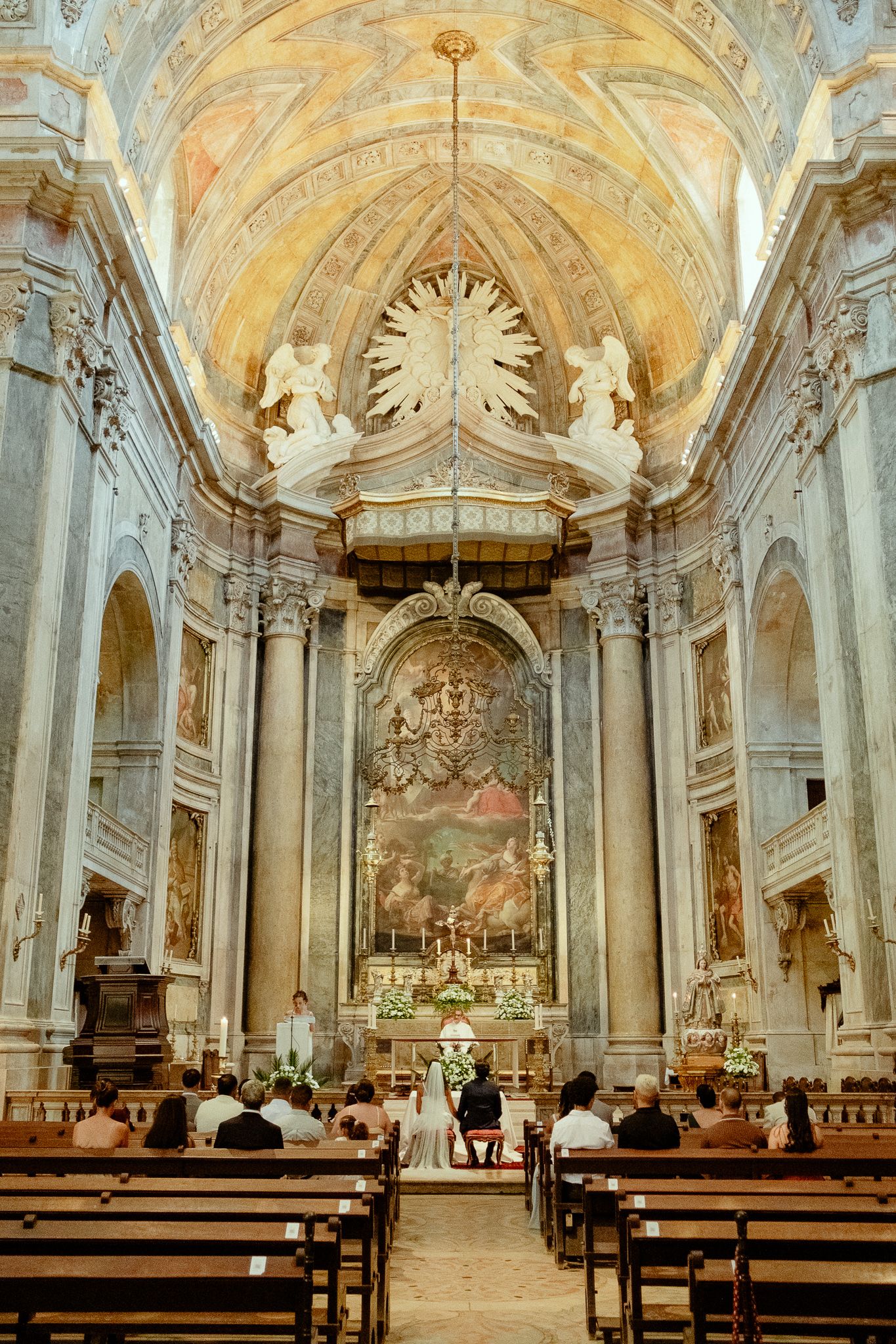 Ornate Baroque church interior with frescoed vaulted ceiling, grand altarpiece painting, and congregation seated in wooden pews.