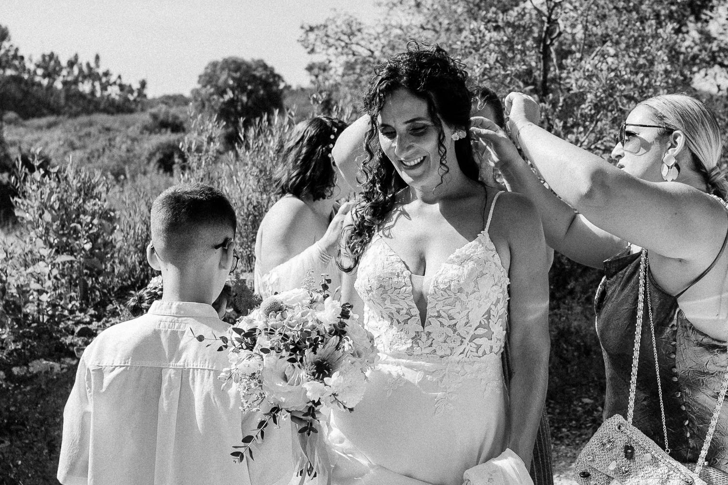 Bride smiling in white lace gown while women adjust her hair outdoors, with a boy holding a bouquet nearby.