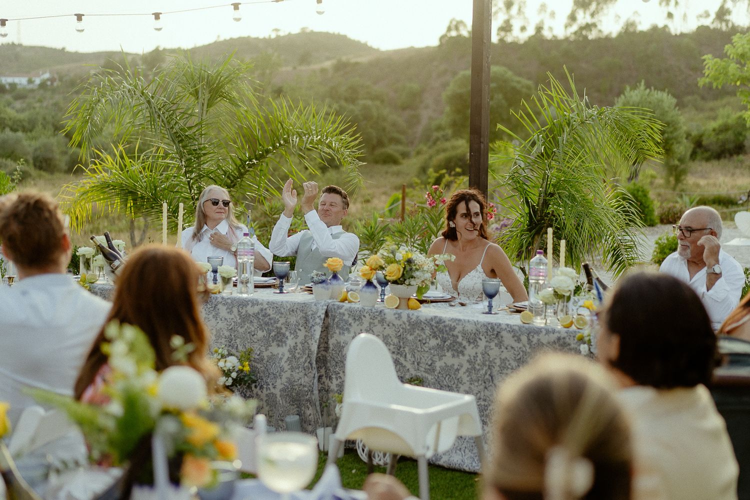 Bride and groom seated at elegant outdoor wedding reception table surrounded by guests and floral arrangements.