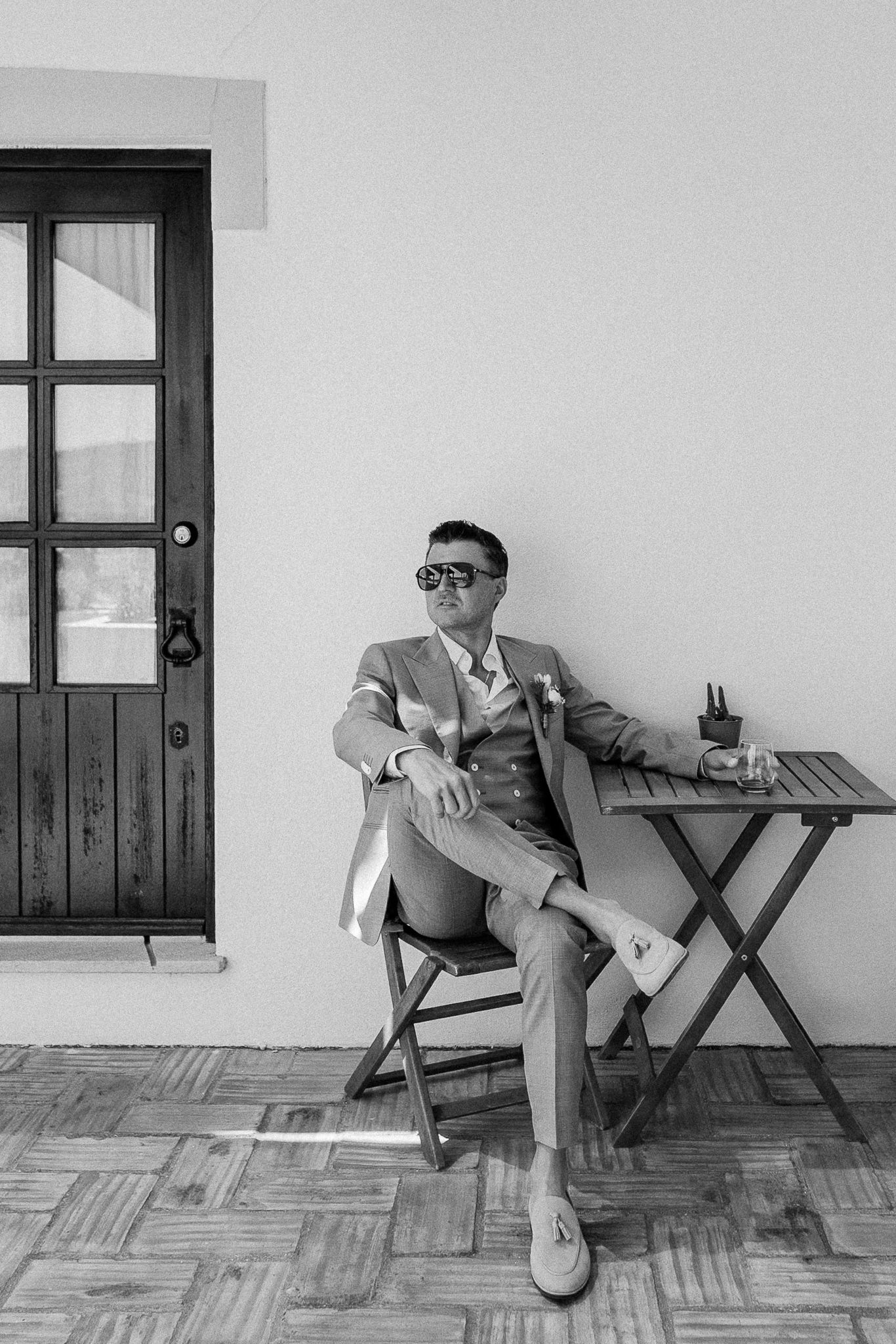 Stylish man in suit and sunglasses sitting at wooden table in black and white photo.