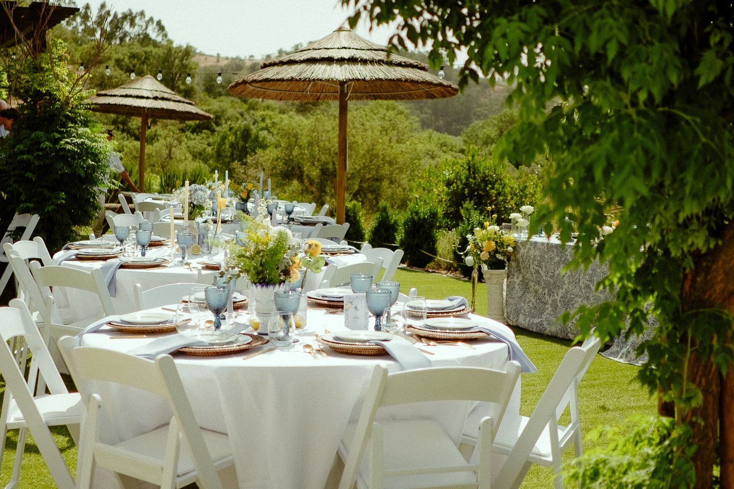 Elegant outdoor wedding reception with white round tables, chairs, and straw umbrellas set on a green lawn.
