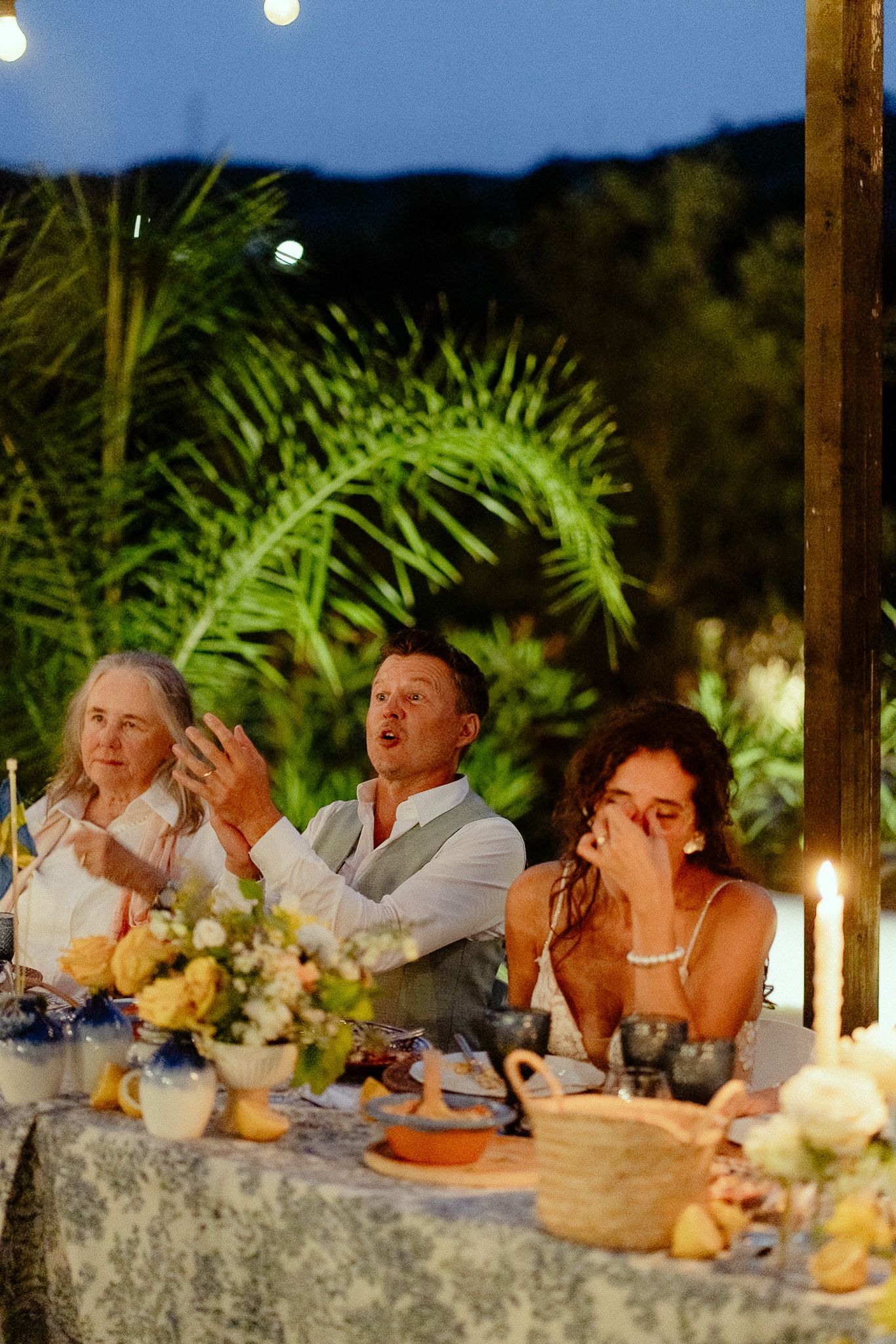 Emotional bride wiping tears at outdoor wedding reception dinner table with candles and floral arrangements at dusk.