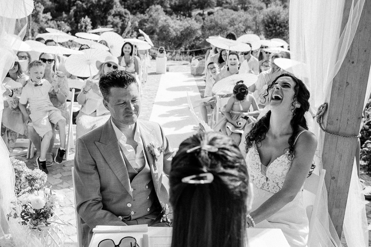 Bride laughing joyfully during outdoor wedding ceremony while groom watches guests seated under sunny sky.