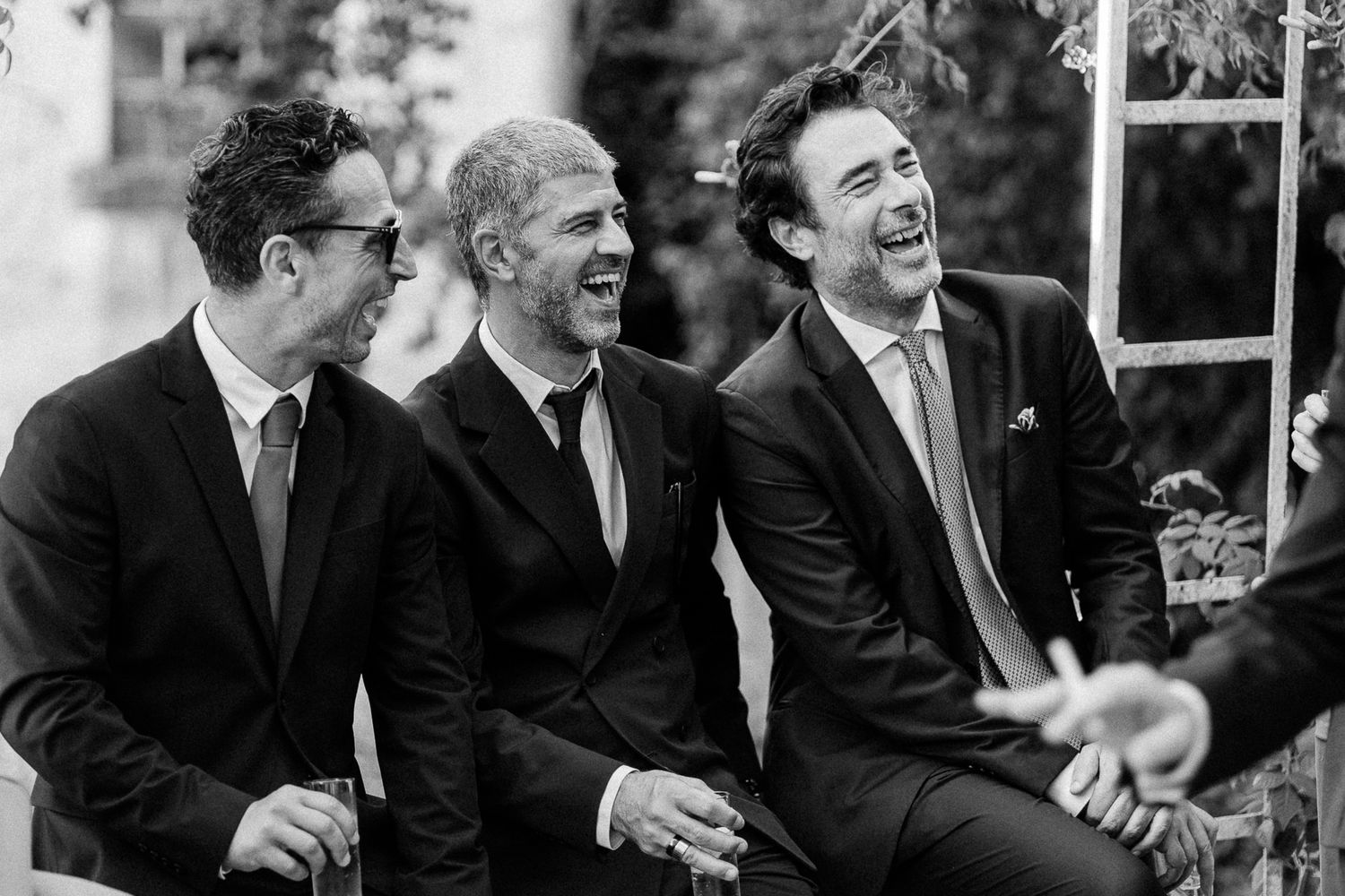 Three men in dark suits laugh joyfully together at a wedding celebration in a black and white photo.