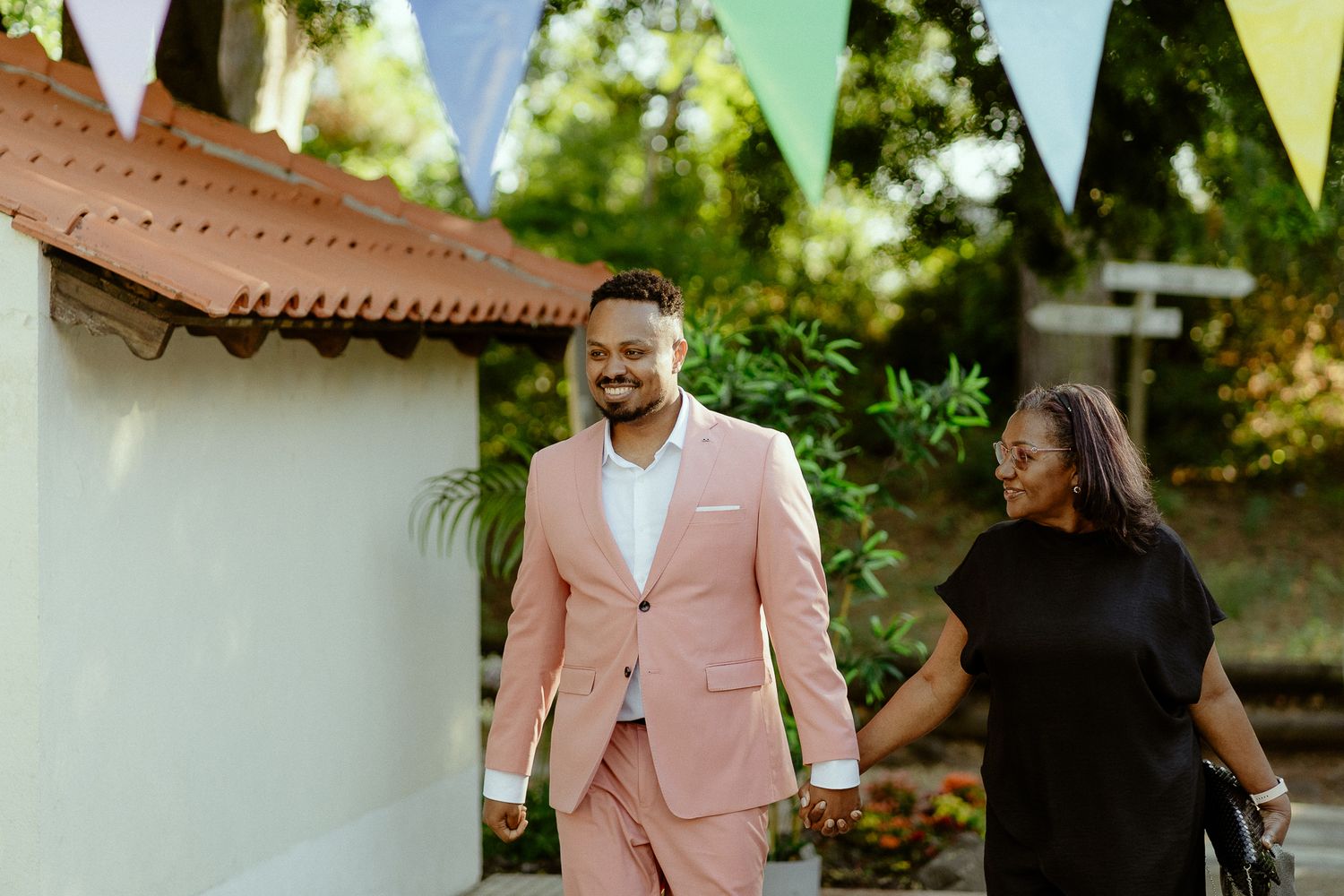 Couple in pink suit and black dress holding hands outdoors at colorful bunting decorated venue.