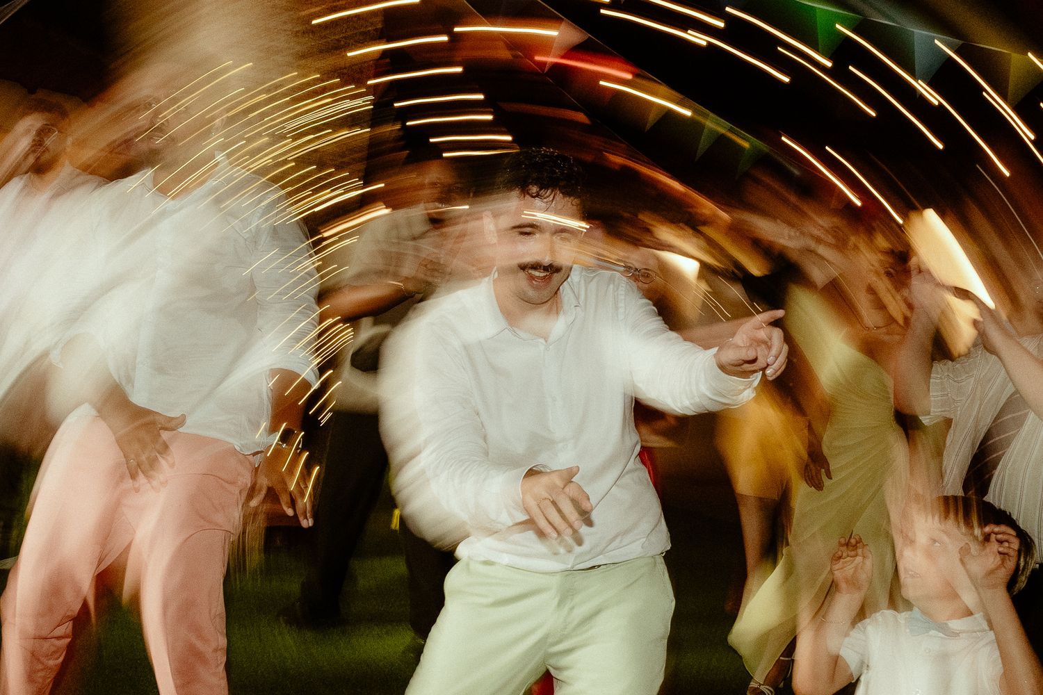 Man in white outfit dancing energetically at an outdoor party with motion blur and colorful lights.