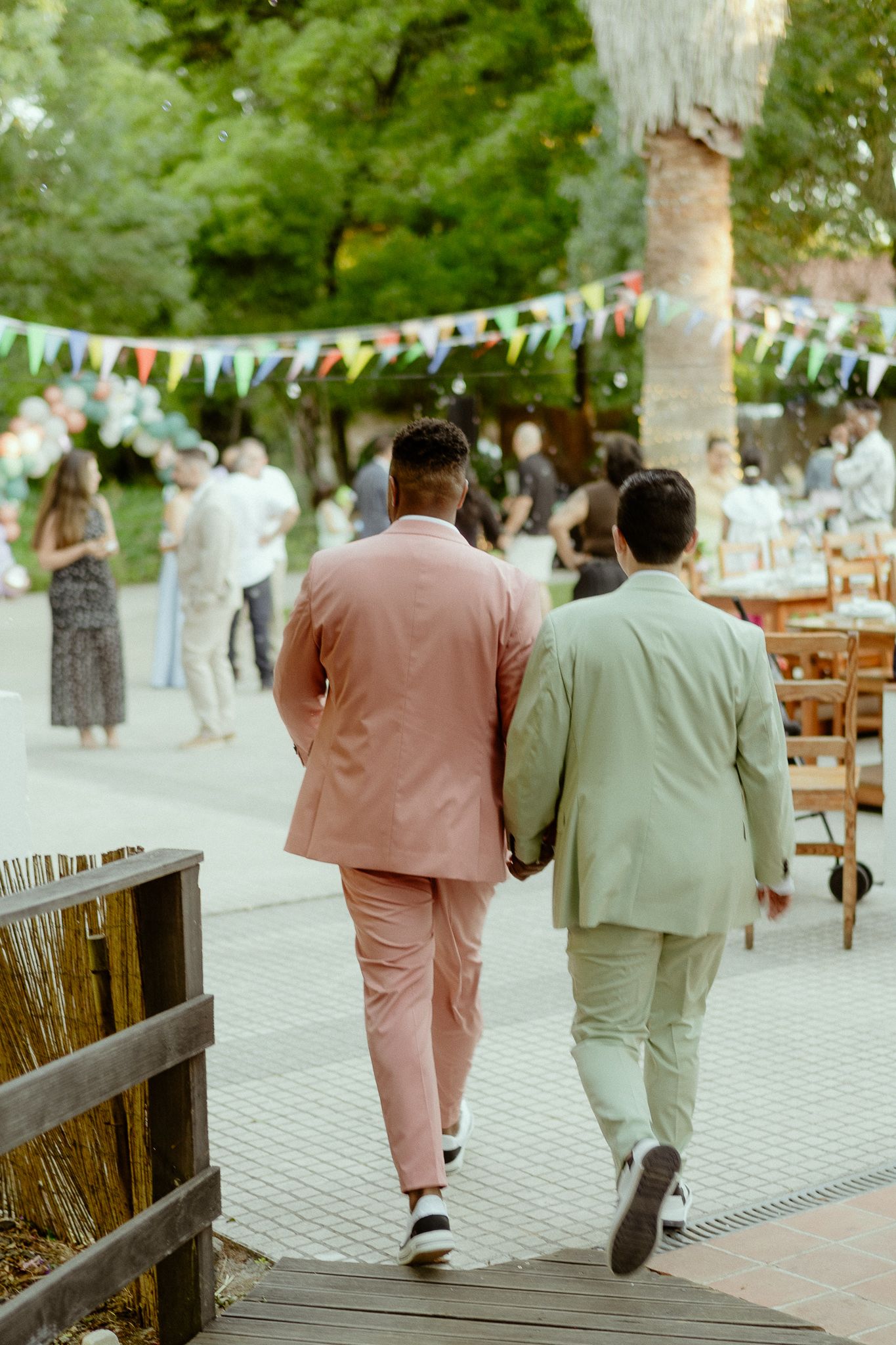 Two grooms in pink and sage suits walk hand-in-hand at an outdoor wedding reception with colorful bunting.