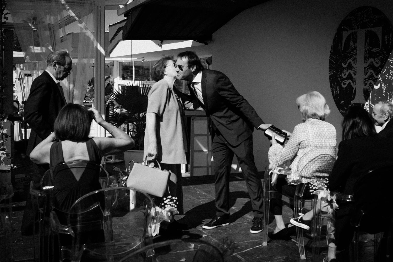 A black and white candid photograph shows people interacting in what appears to be a busy indoor social gathering.
