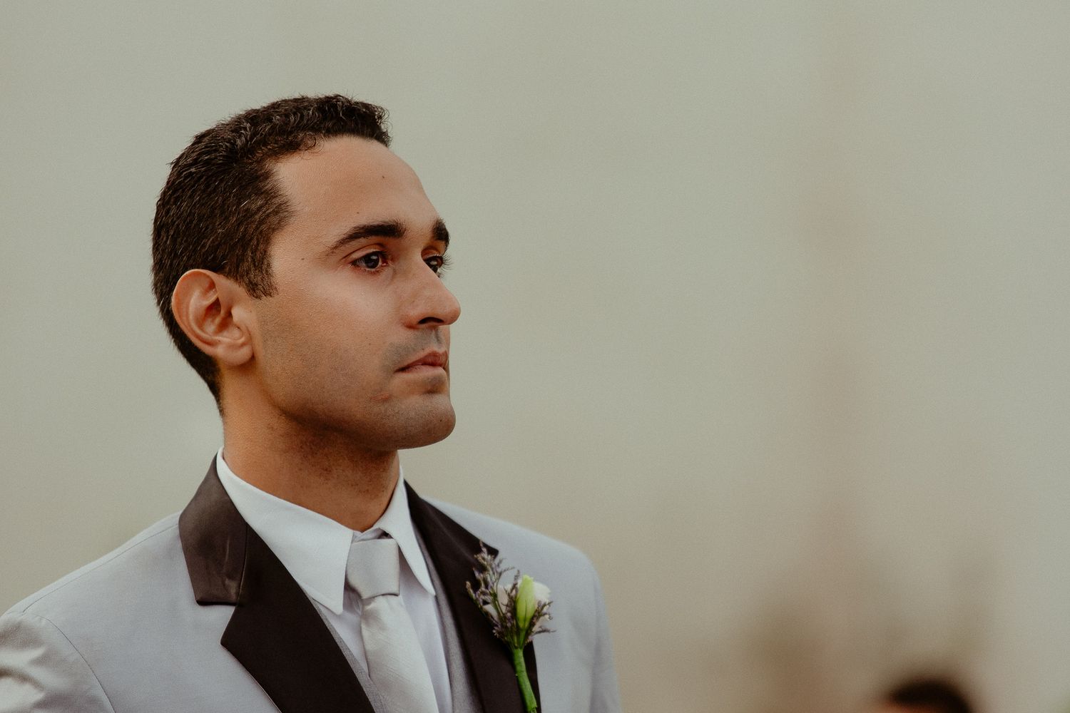 A person in a gray suit jacket with a boutonniere looks to the side against a blurred background.