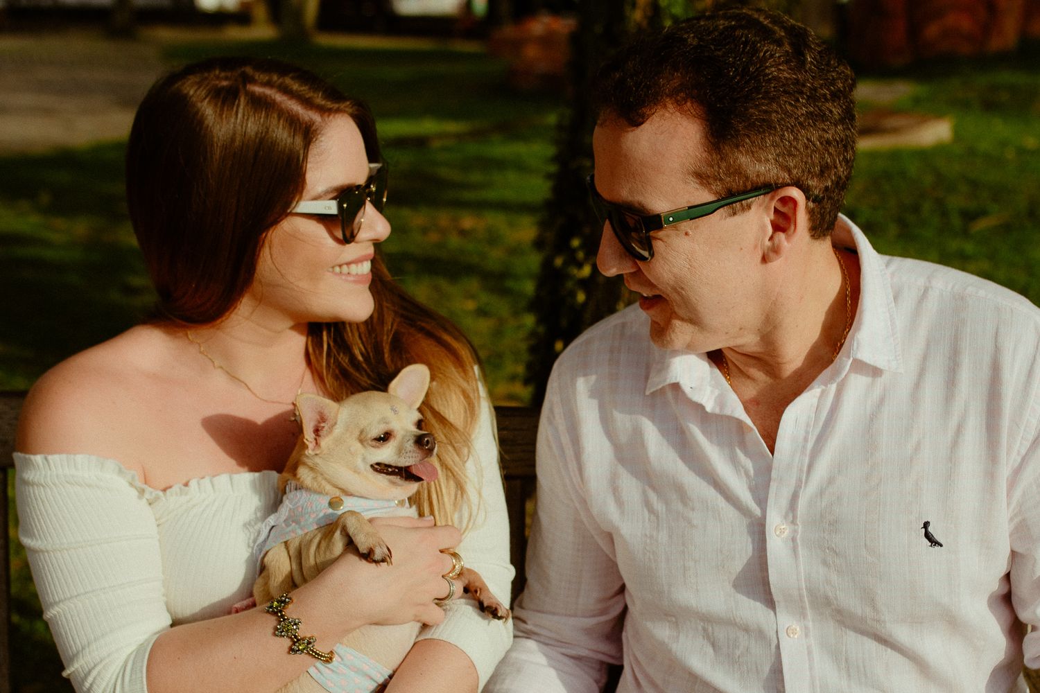 A couple in sunglasses sitting outdoors while holding a small white cat in their arms.