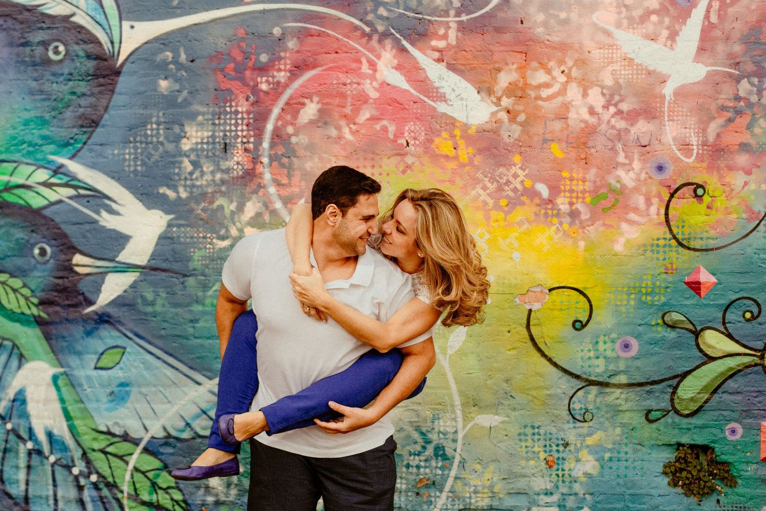 Couple dancing gracefully against a vibrant mural background with birds and swirling artistic elements.