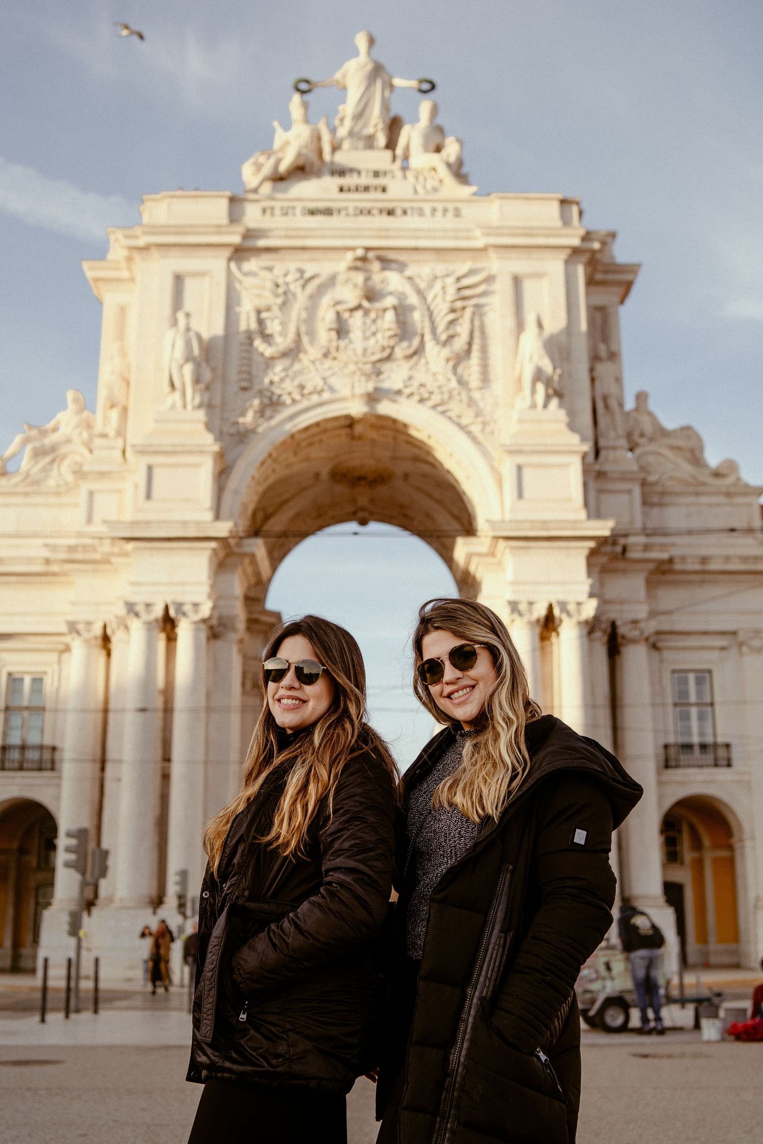 Tourists posing in front of ornate white stone arch at Comercio Square in Lisbon.