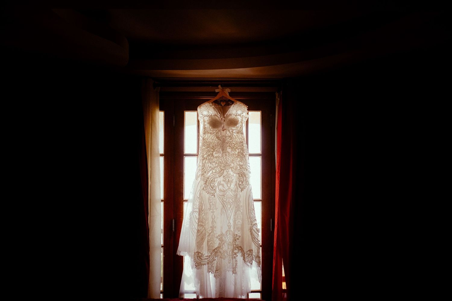 A white lace wedding dress hangs silhouetted against a sunlit window with red curtains in a dark room.