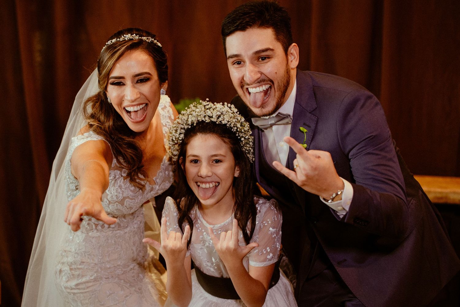 A joyful wedding celebration with a bride, groom and flower girl showing excitement and laughter in an indoor setting.
