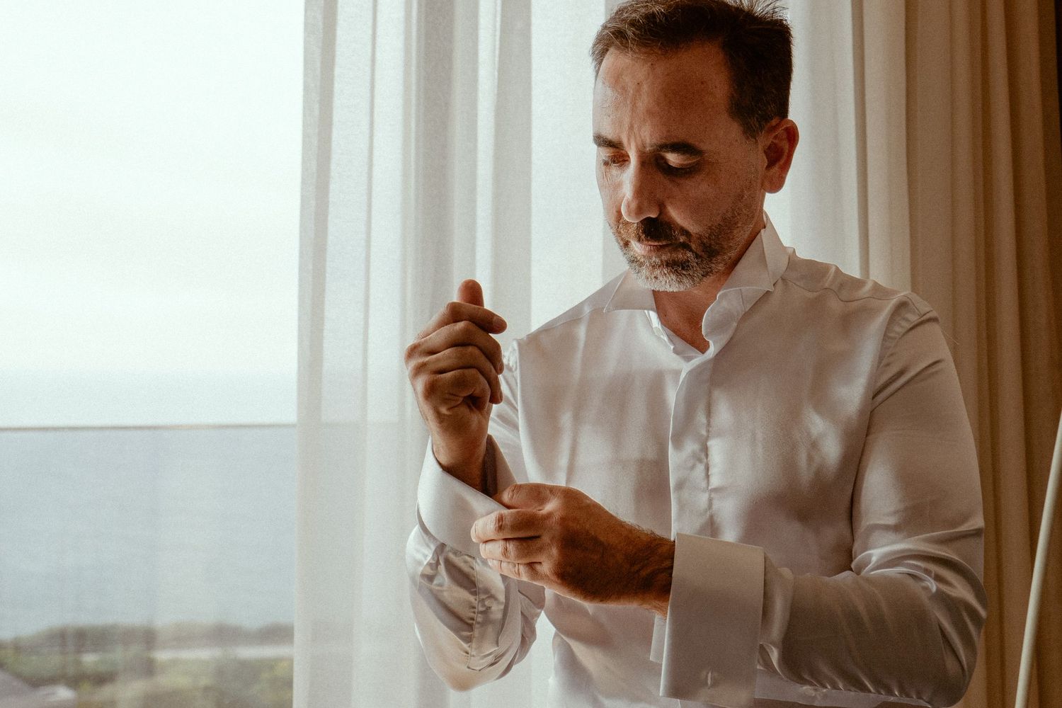 Man in white dress shirt adjusts his tie while standing in front of a bright window.