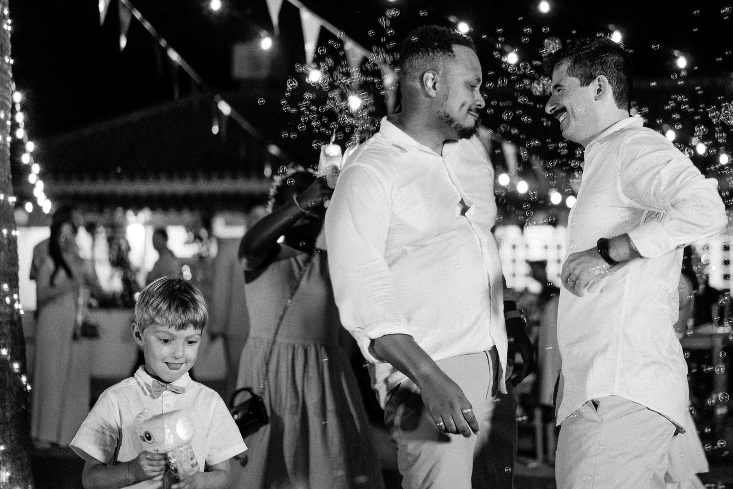 Black and white photo of two men in white shirts celebrating at an outdoor evening party with string lights and a child nearby.