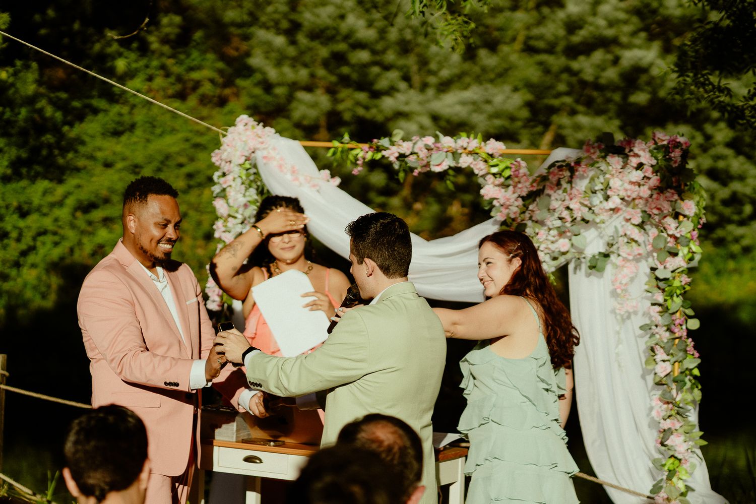 Outdoor wedding ceremony with groom in pink suit, officiant, and bridesmaid under floral arch.