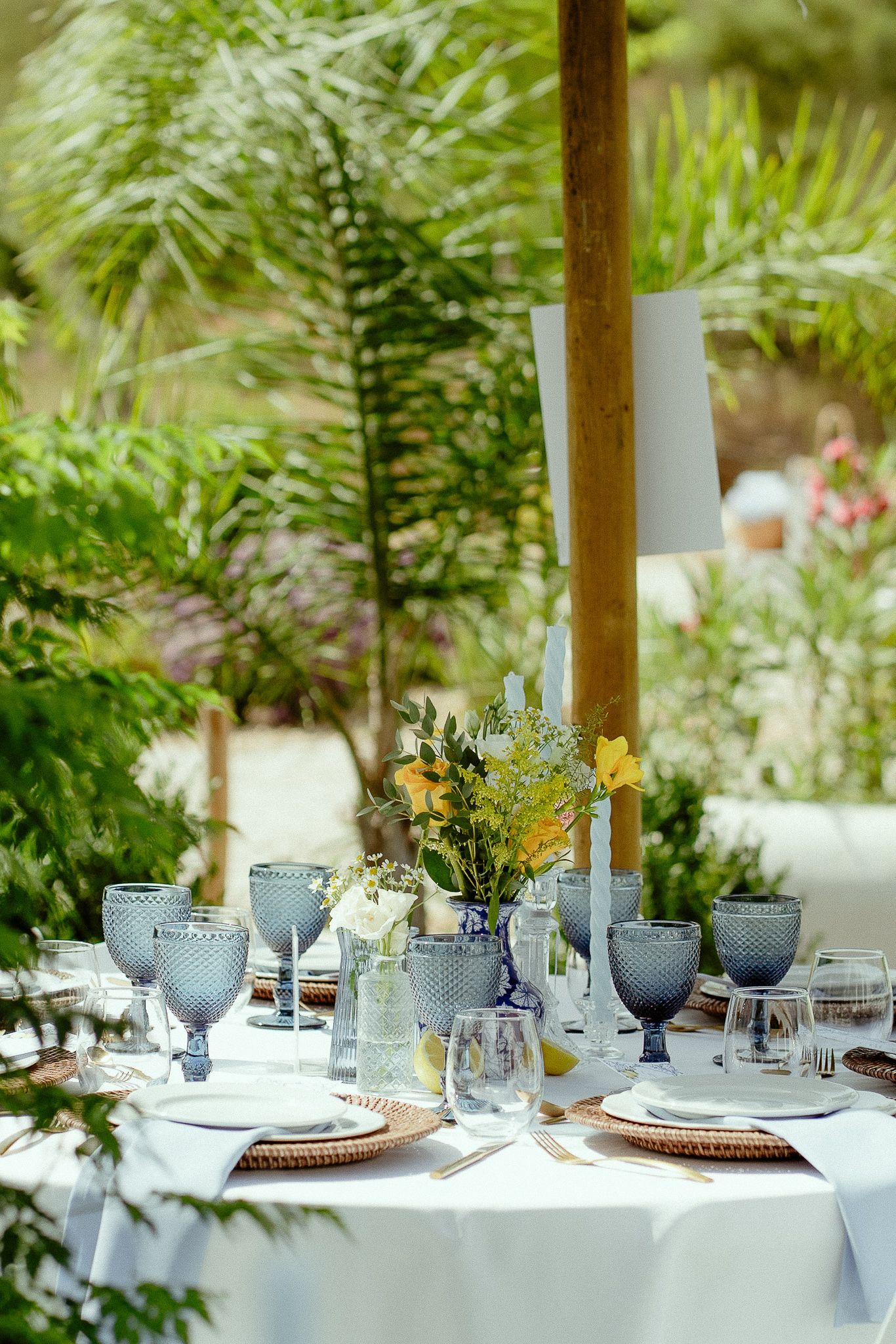Elegant outdoor dining table set with blue glassware, floral centerpiece, and white linens surrounded by lush greenery.