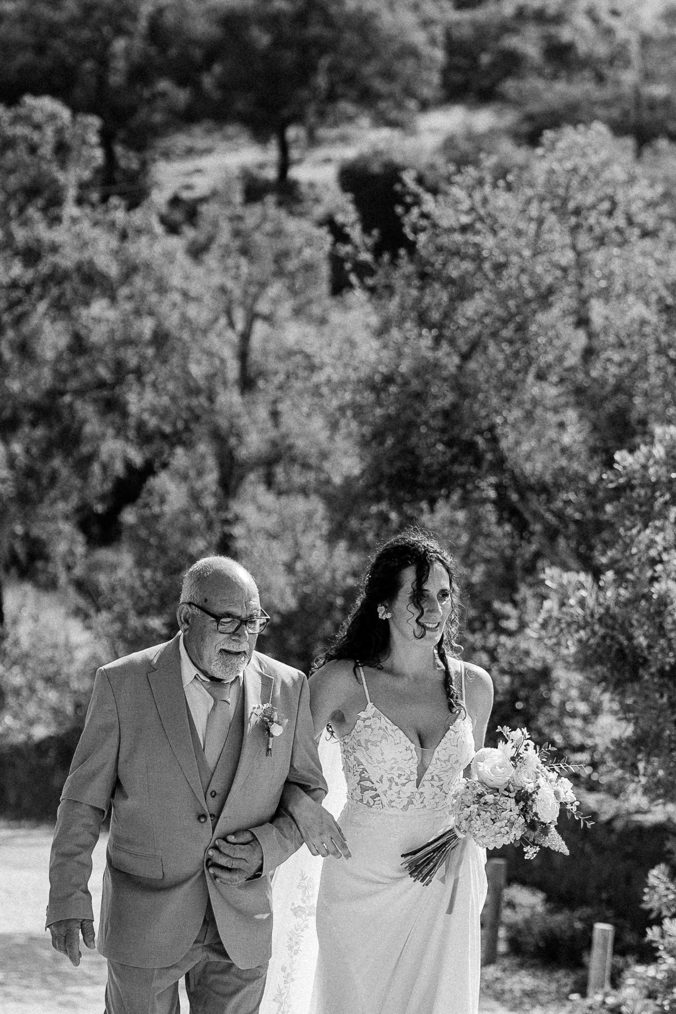 Black and white photo of a bride holding bouquet walking arm in arm with older man in suit at outdoor wedding.