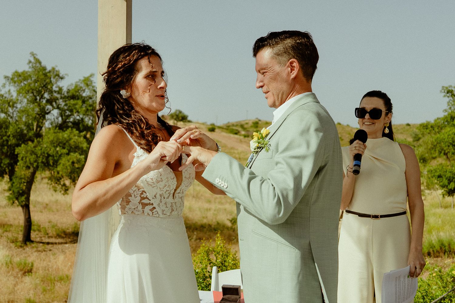 Bride and groom exchange rings during outdoor wedding ceremony, officiant holding microphone in background.