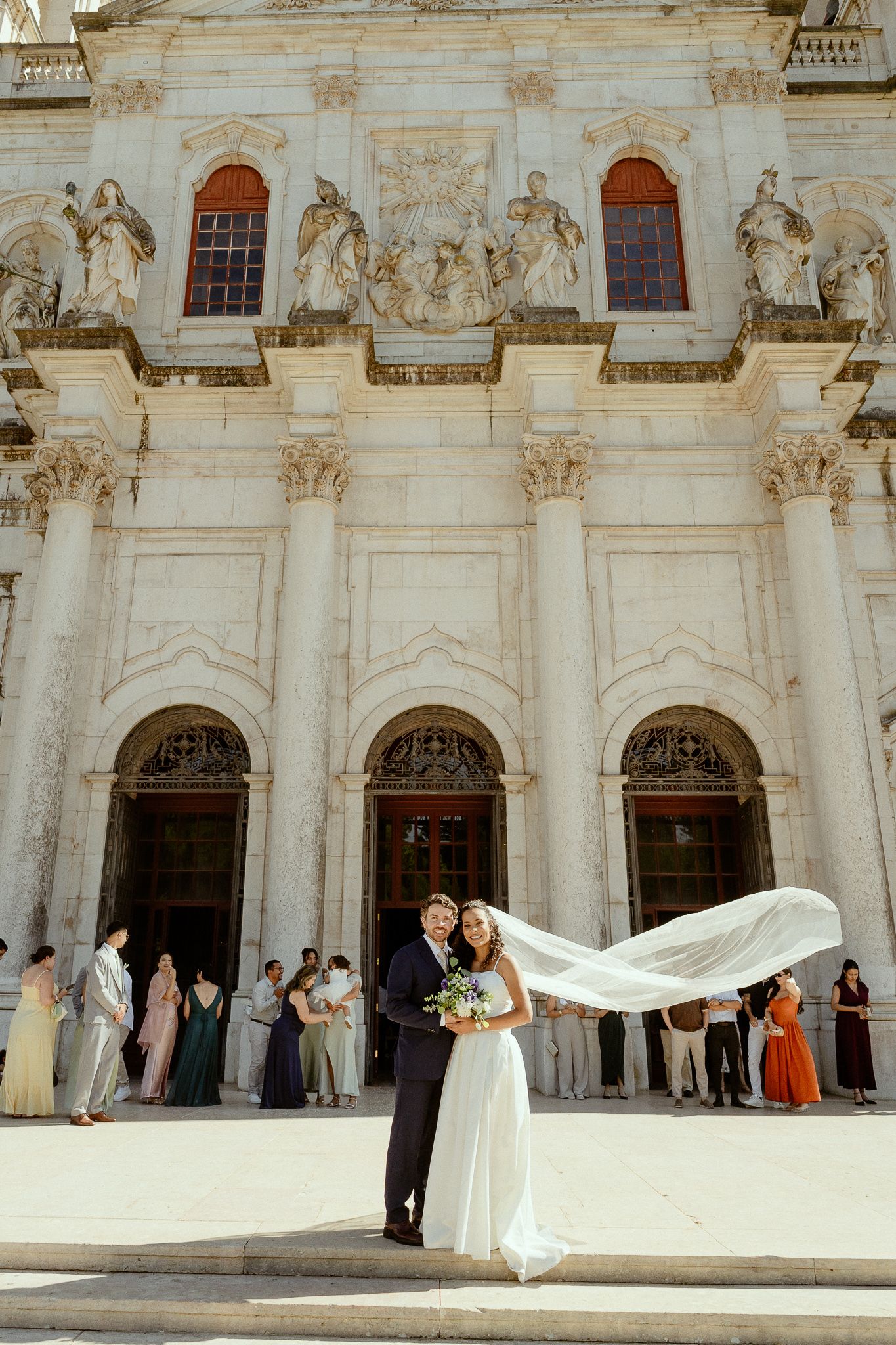 Bride and groom pose before grand white baroque church facade, veil flowing in wind, wedding guests gathered behind them.
